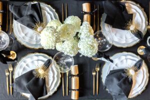 Vogelperspektive eines festlich gedeckten Silvester-Tisches in Schwarz, Gold und Weiß mit eleganten Details wie Knallbonbons, Chrysanthemen und goldenem Besteck.
