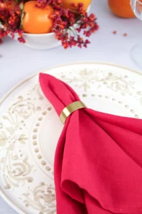 Close-up of a gold napkin ring with hand-painted plate from the Adelasia collection and cherry-red linen napkin and gold-coloured napkin ring.