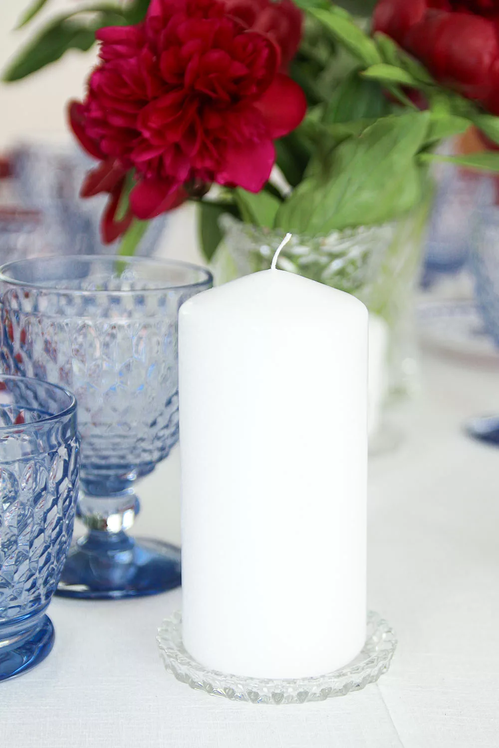 magnolias-on-silk-table-of-the-month-mai-peony-red-blue-table-inspiration-24 White pillar candle on a glass coaster between blue water glasses and red peonies.