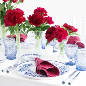 Close-up of a laid plate with red linen napkin, blue and white decorative plate and crystal glasses on a white tablecloth with peonies in the background.