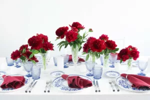 Generously laid table with red peonies, blue glasses and blue and white crockery on a white tablecloth.
