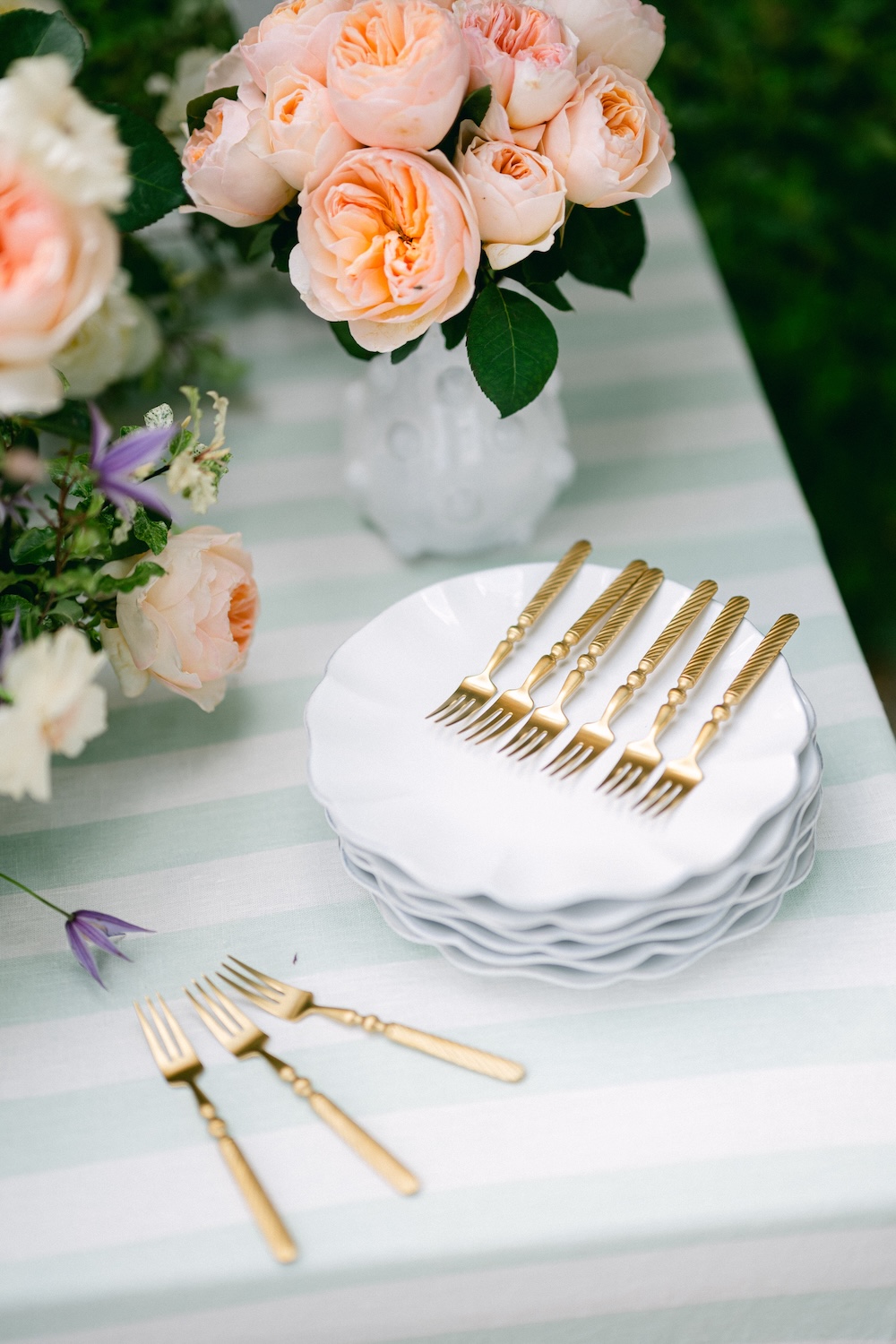 Designed summer table with peach roses, green and white striped tablecloth, stacks of white plates and golden dessert forks in Magnolias on Silk style.