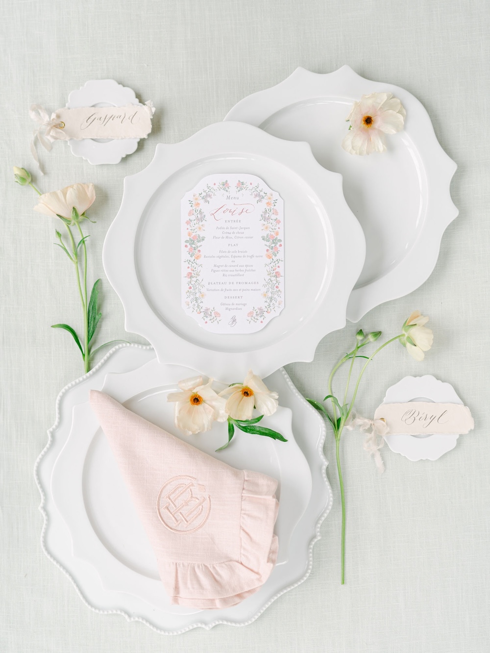 Flaches Flatlay eines eleganten Place Settings mit weißen Barocktellern, floraler Menukarte, pastelligen Blüten und roséfarbener Rüschenserviette – inspiriert von französischer Belle-Epoque-Ästhetik.