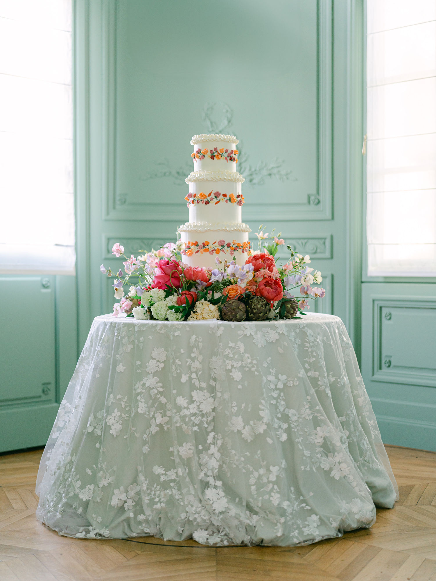 Four-tier wedding cake on a floral base, presented on a draped lace tablecloth in a pastel green parlour - an example of layering and textured table design.