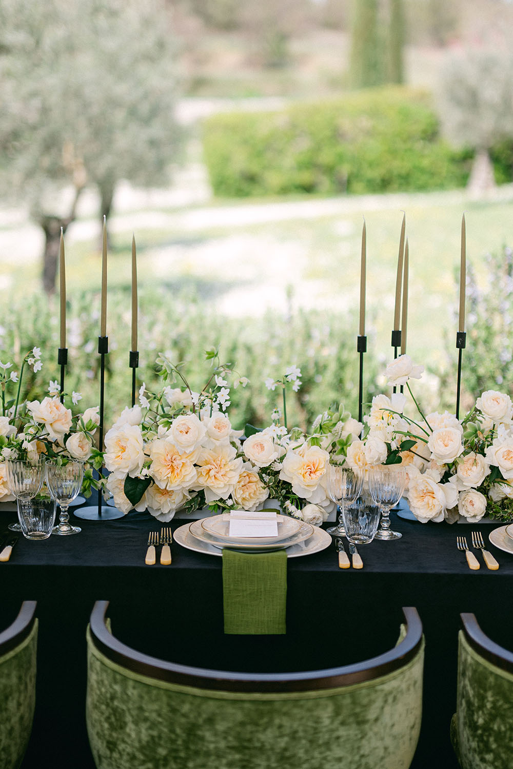 Long table with cream-coloured roses, dark candlesticks and green napkins in an outdoor setting.