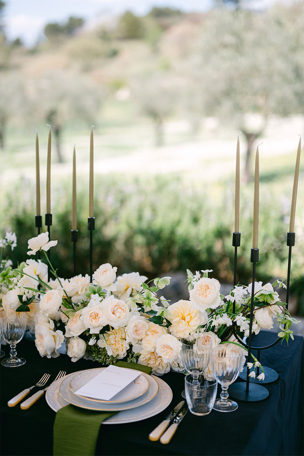 Elegantes Outdoor-Tischsetting mit cremefarbenen Rosen, moosgrüner Serviette und schwarzen Kerzenhaltern.